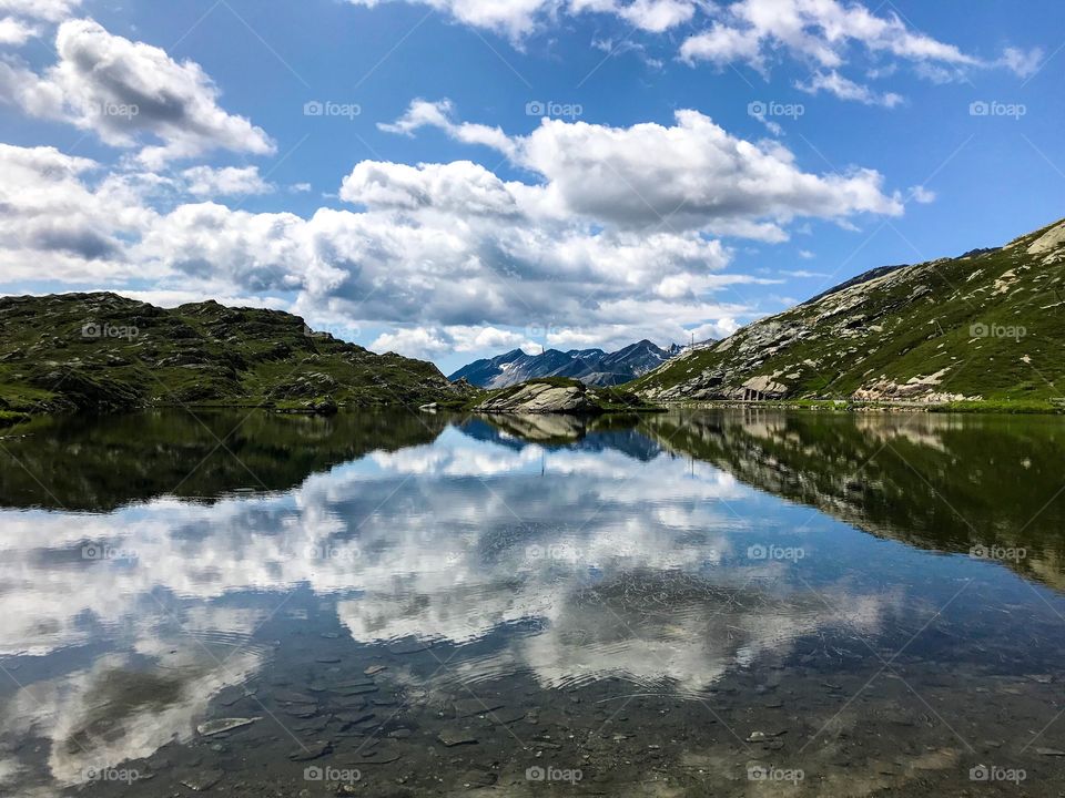 a lake in the middle of nature, with mountains surrounding it, clear water visible can on the bottom of the lake. in the blue sky there are large white clouds, color reflection  into the water looks amazing