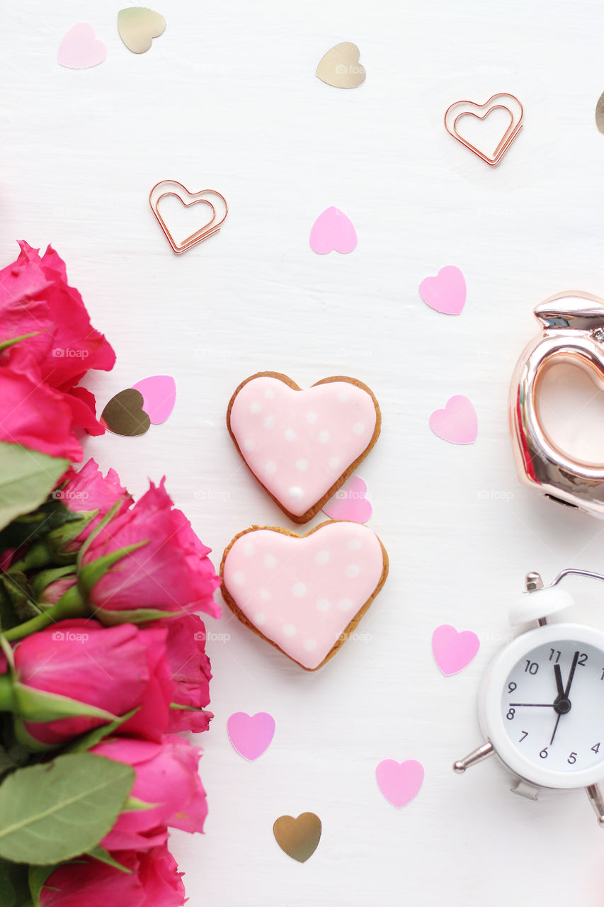 two gingerbread hearts, a bouquet of bright pink roses, an alarm clock and plastic hearts on a white table. flat lay