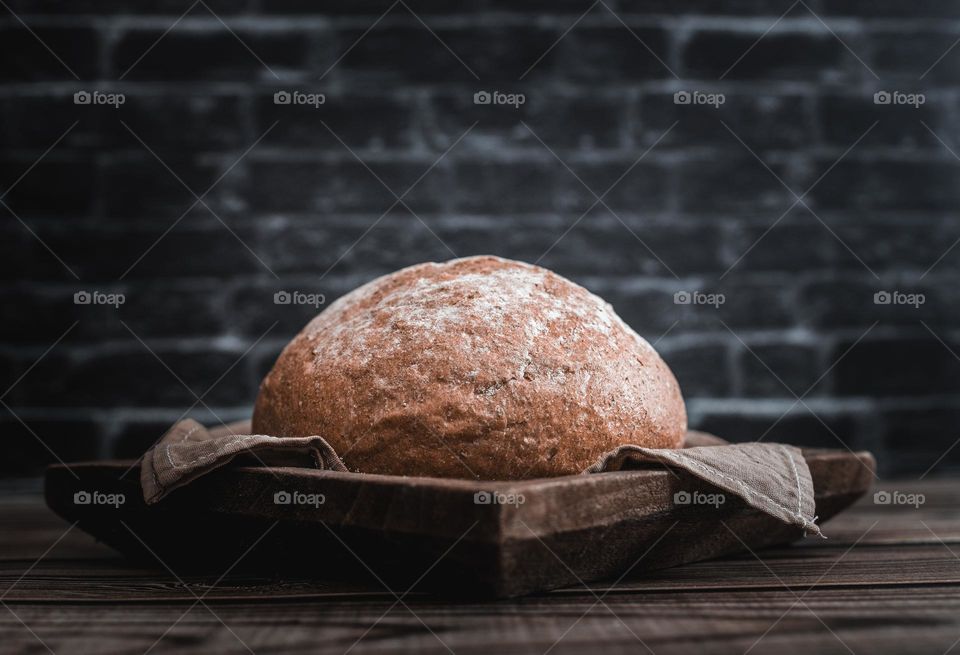 One round rye bread with cereals fiber in a wooden brown square plate with a cotton napkin lies in the center on the table against the background of a black blurred brick wall, close-up side view. The concept of baking bread.
