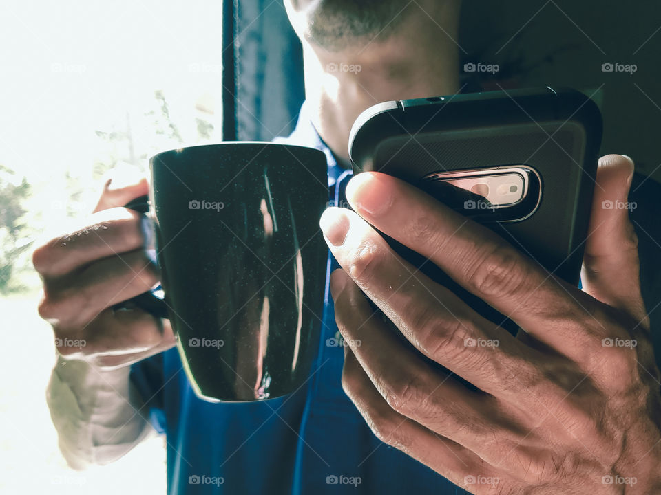 Closeup of a man drinking coffee by the window, while reading on his phone and drinking out of his favorite gun metal gray, fire glazed, ceramic coffee mug.