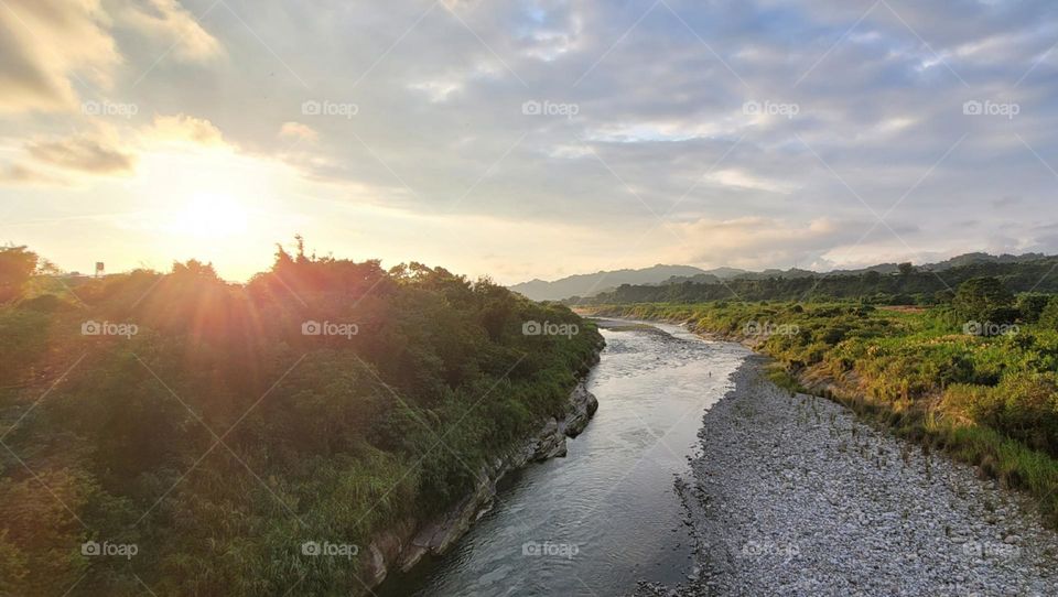 The sun shines through the clouds on the mountains and the stream.