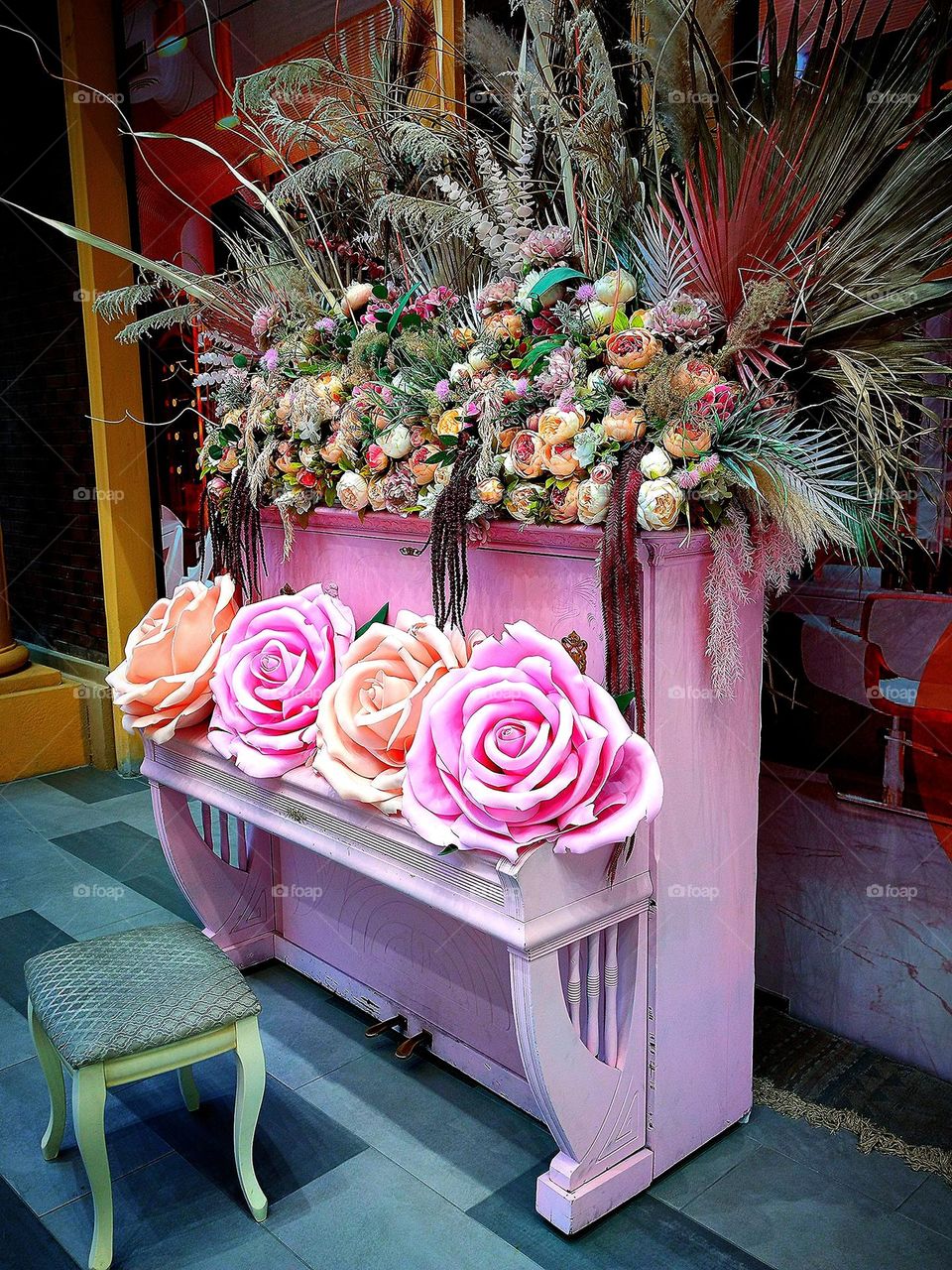 Baby pink.  A baby pink piano with artificial roses in different shades of pink on the lid.  On the top of the piano lie multi-colored flowers and ears of cereals.