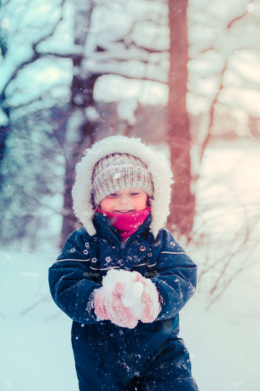 Happy smiling little girl enjoying snow. Toddler is playing outdoors in wintertime while snow falling. Toddler is wearing dark blue snowsuit and wool cap