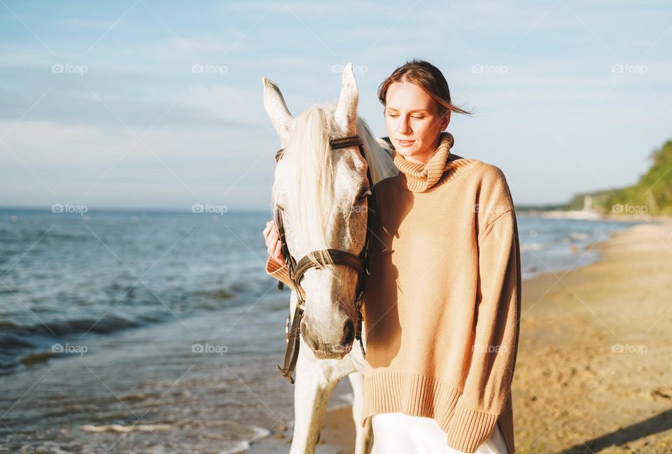 Young long hair woman in beige sweater with white horse on seascape background