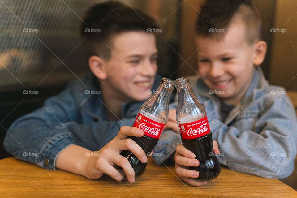 two lovely brothers, dressed in denim clothes, with beautiful hairstyles, are sitting in a cafe and drinking Coca-Cola.  communicate, laugh, smile.  two guys, two friends