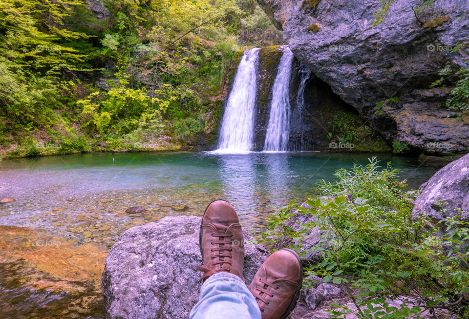 Feet in brown shoes sitting on the rock by the lake and waterfall in front of a gorgeous mountain landscape at Olympus mountain in Greece.