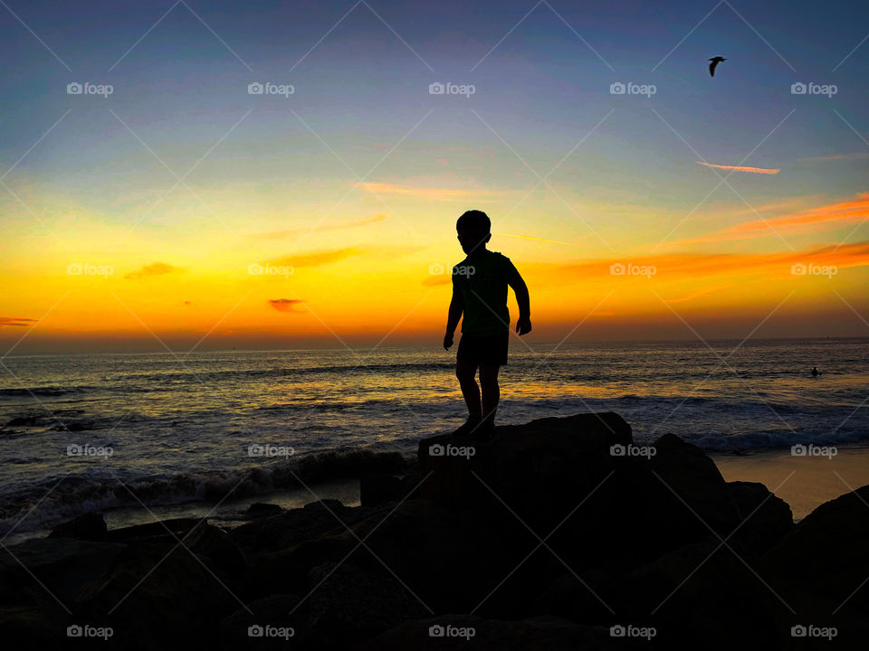 Silhouette of boy at a California beach during sunset. The stark black silhouette contrasts with the bright blue and orange sunset.