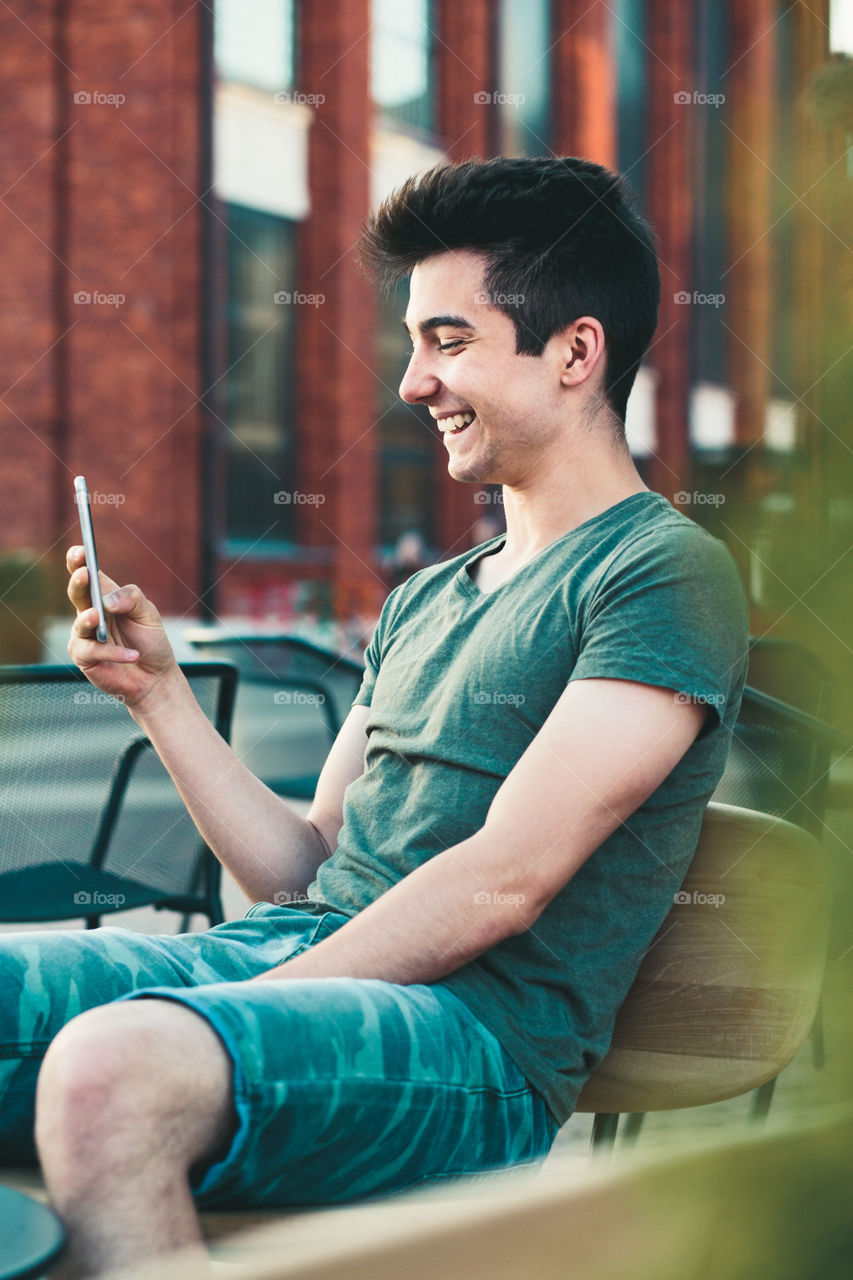 Young man having fun with smartphone, reading funny texts, sitting in center of town