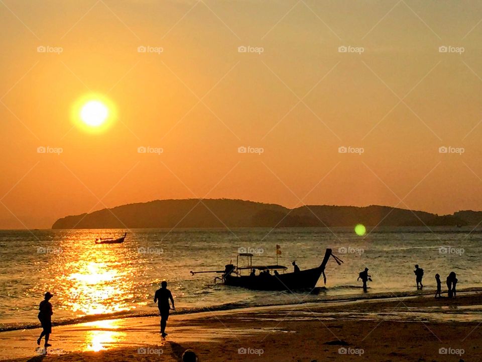 Stunning magic dramatic sunset over the smooth sea water surface with the islands at the horizon and the dark silhouette of the fisherman longtail boat and people on the shore beach