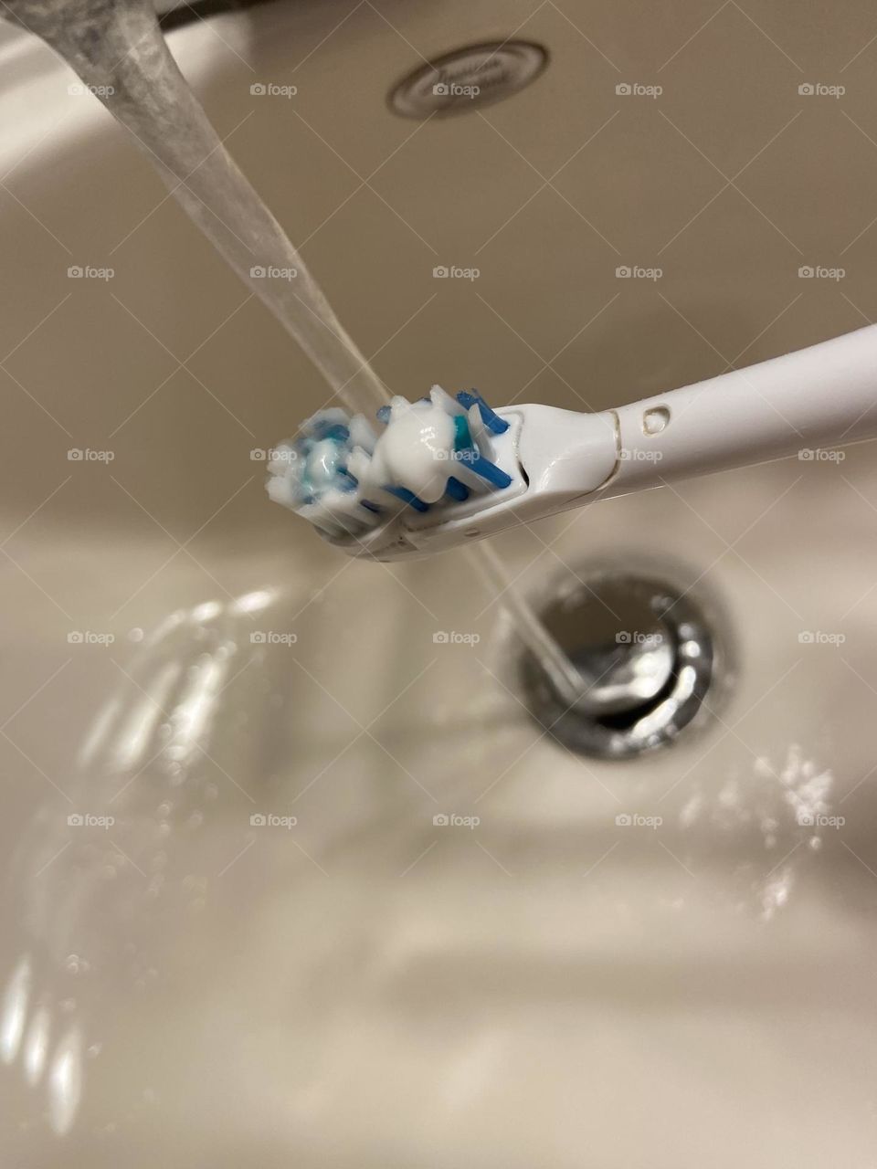Wetting my electric toothbrush with water from the faucet in the bathroom sink in preparation for brushing my teeth 