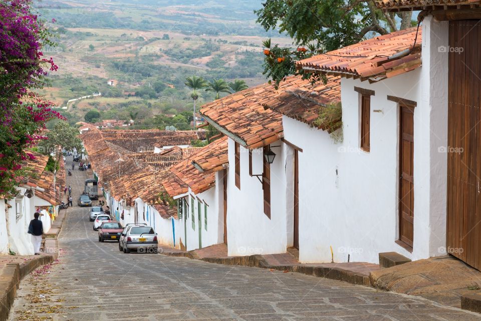 Village with red tiled roofs. Small downhill village road. White houses next to the street. Red tiled roofs on houses. Few cars parked.