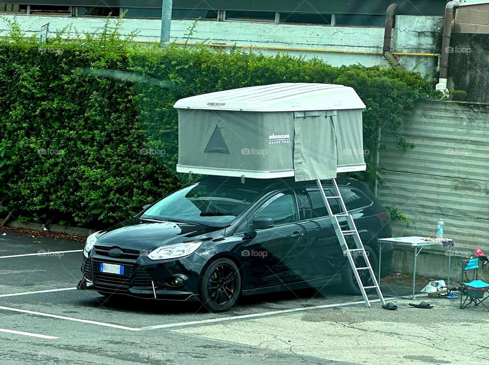 Summer in the city.  There is a car in the parking lot, on the roof of which there is a tent.  There is a ladder attached to the tent.  There is a folding table next to the car.  Summer shoes on the pavement