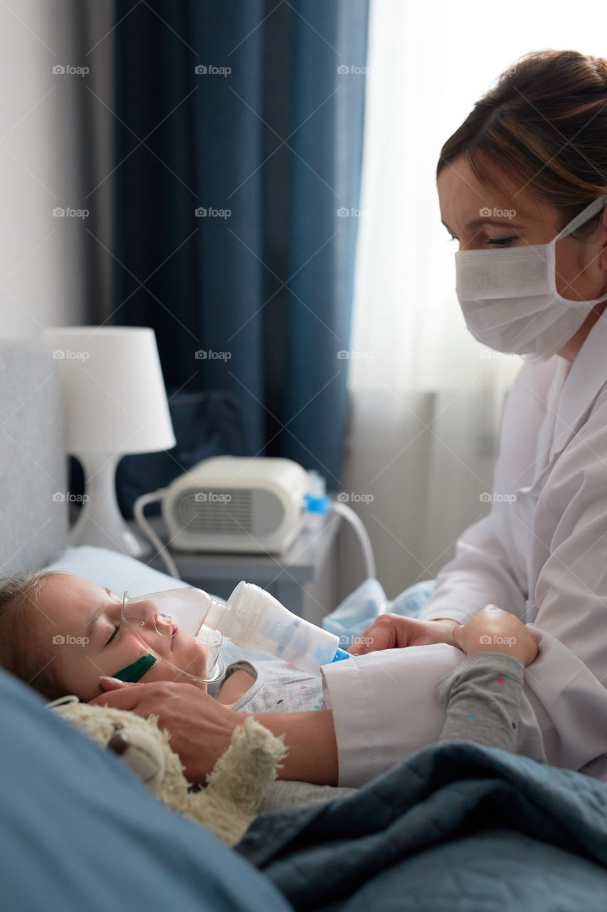 Doctor visiting little patient at home. Child having medical inhalation treatment with nebuliser. Woman wearing uniform and face mask