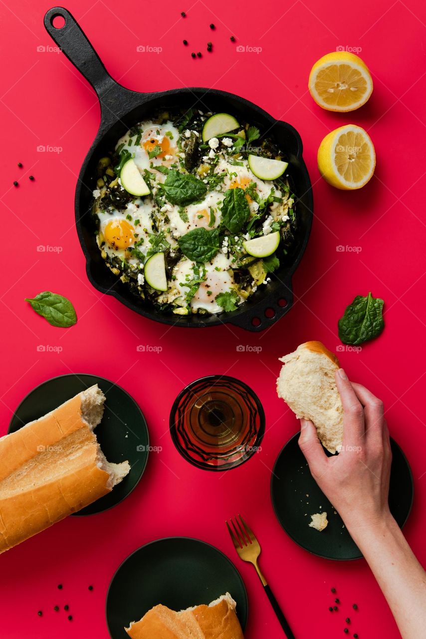 Colorful flat lay of a green shakshuka skillet dish with a person’s hand holding a torn piece of bread loaf