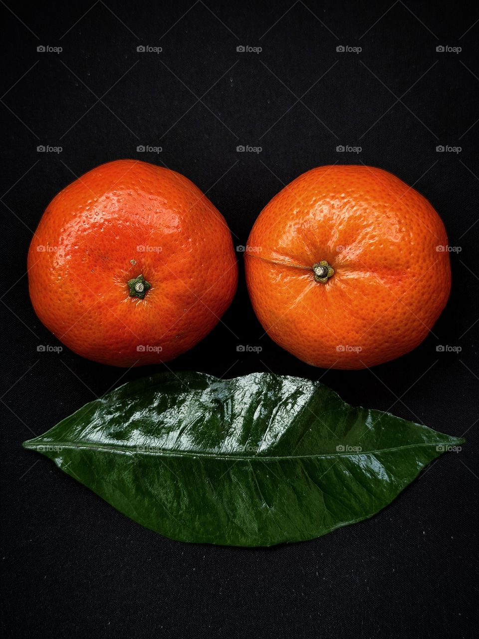 Two orange tangerines and below them a green tangerine leaf.  the composition resembles two eyes and a mouth.  On a black background