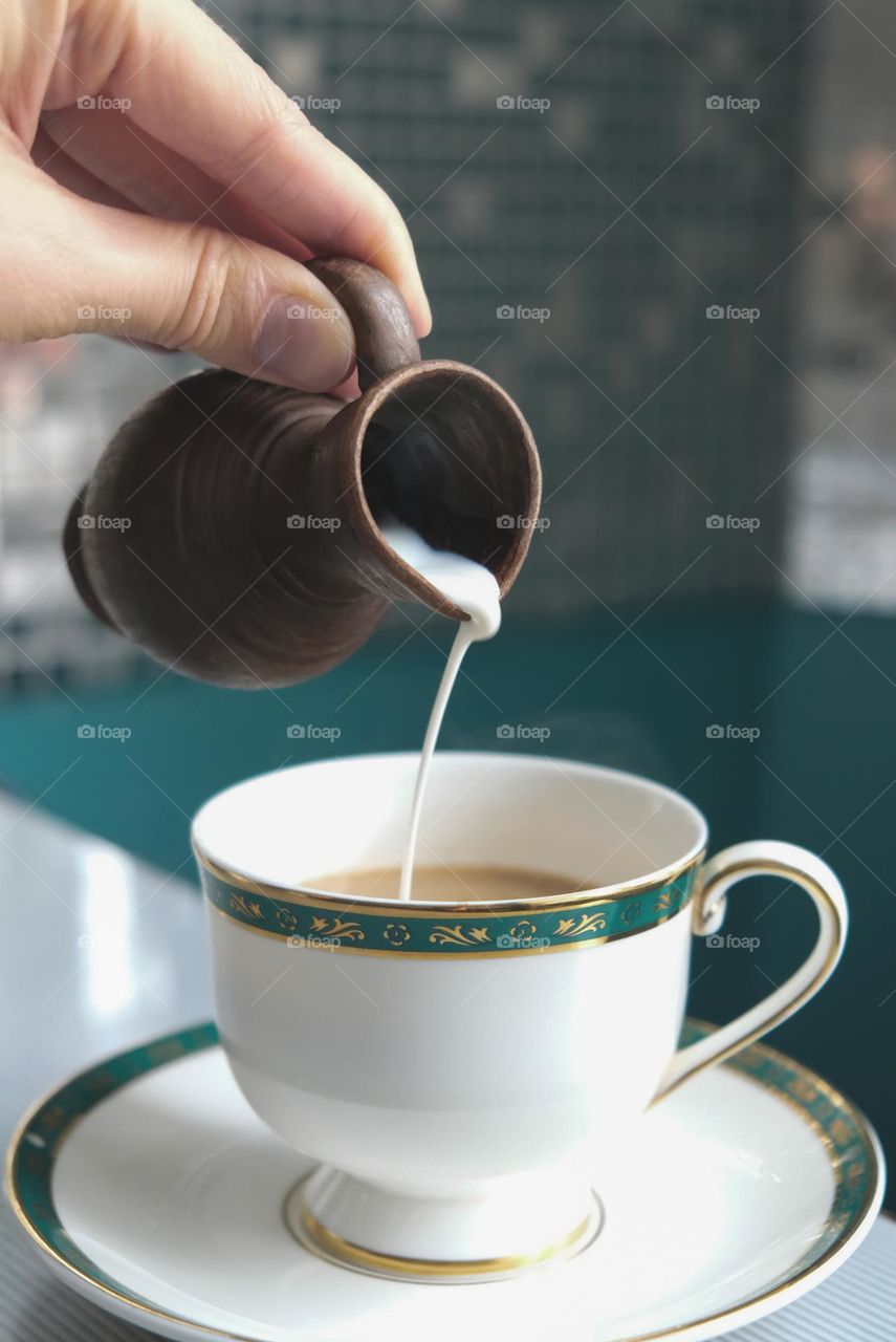 Hand pours milk from a clay milk jug into a cup of coffee