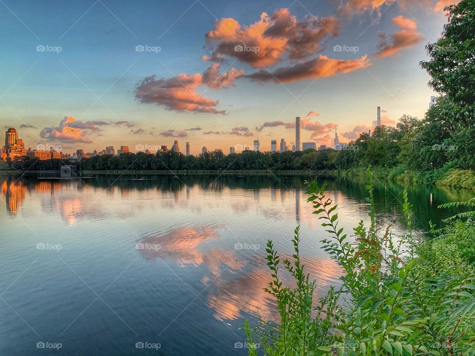 Beautiful reflections of blue sky, orange clouds, green trees, building in the reservoir in New York.