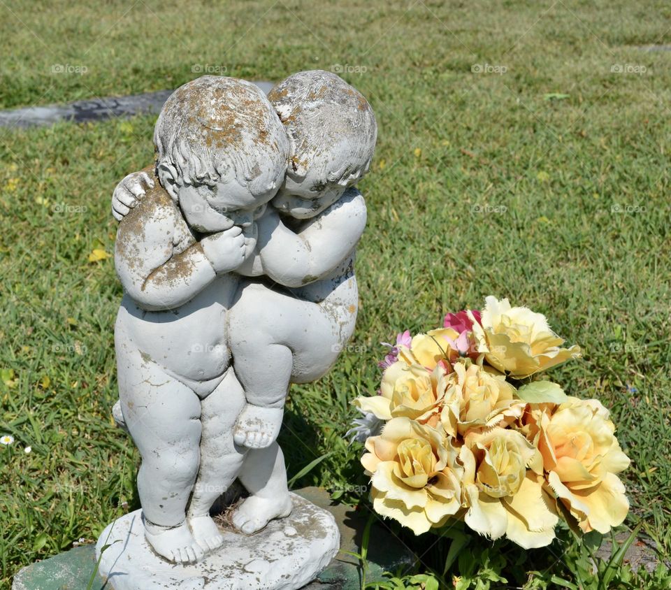 A sculpture of two toddlers hugging standing next to faded yellow fake flowers outside on the grass