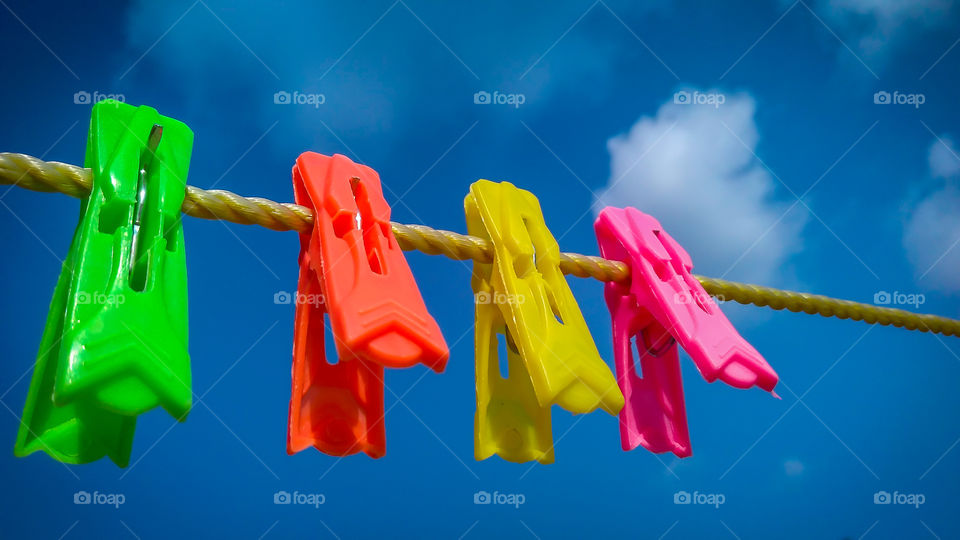 A group of colorful clips hanged together in a yellow rope behind a blue sky with white clouds.