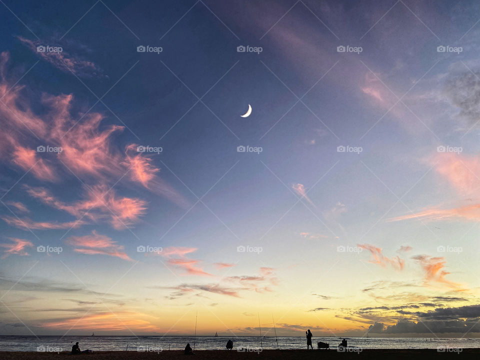 A crescent moon over silhouetted figures fishing on a beach at sunset