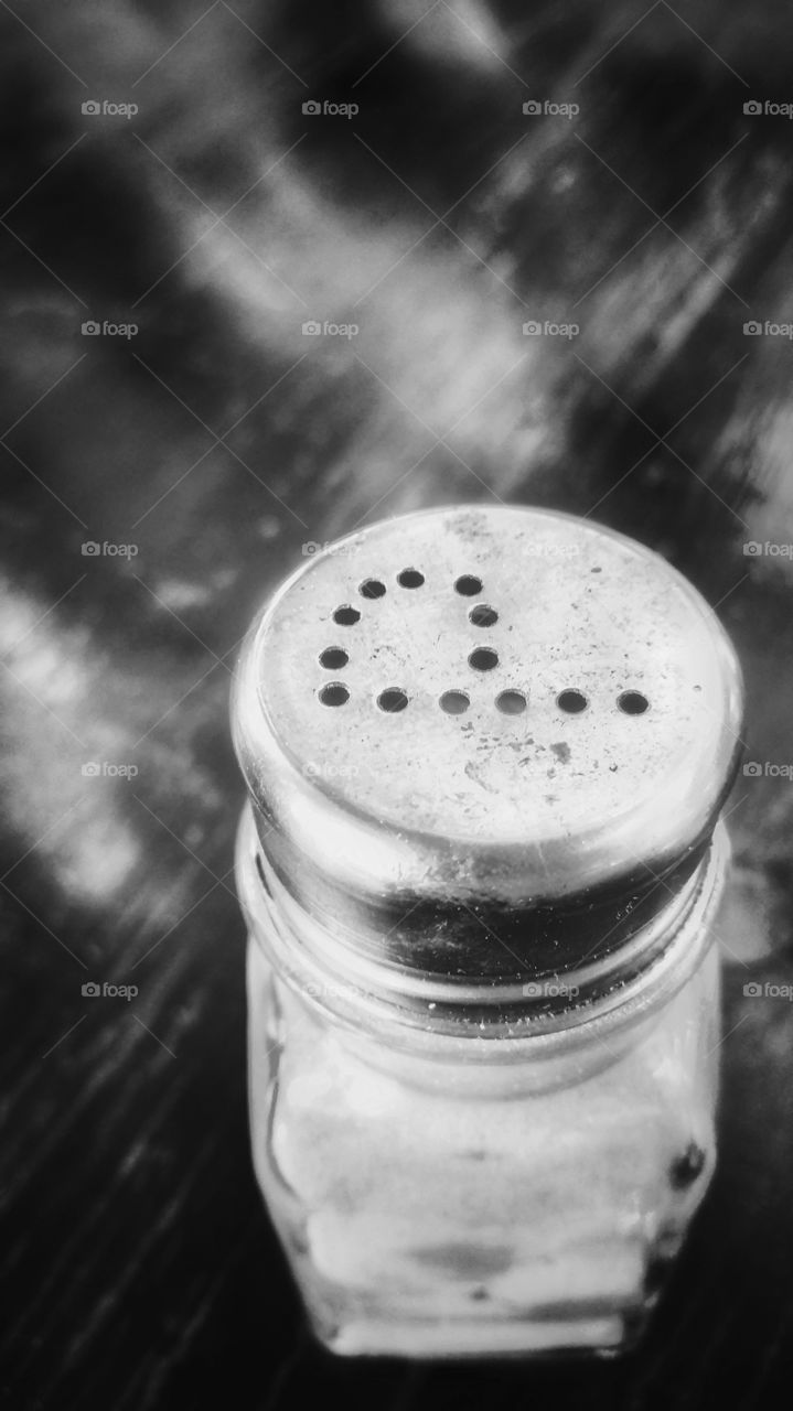 A pepper shaker on a wooden table with light reflections. Small round dots punched to the top of the shaker confirms it's content. Monochrome image.