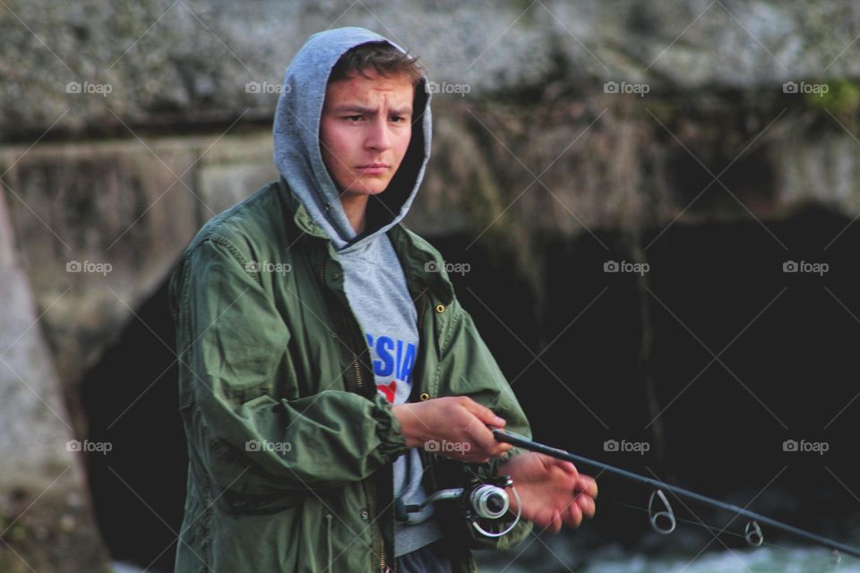 a fisherman in a green cloak and hood holds a spinning rod in his hands