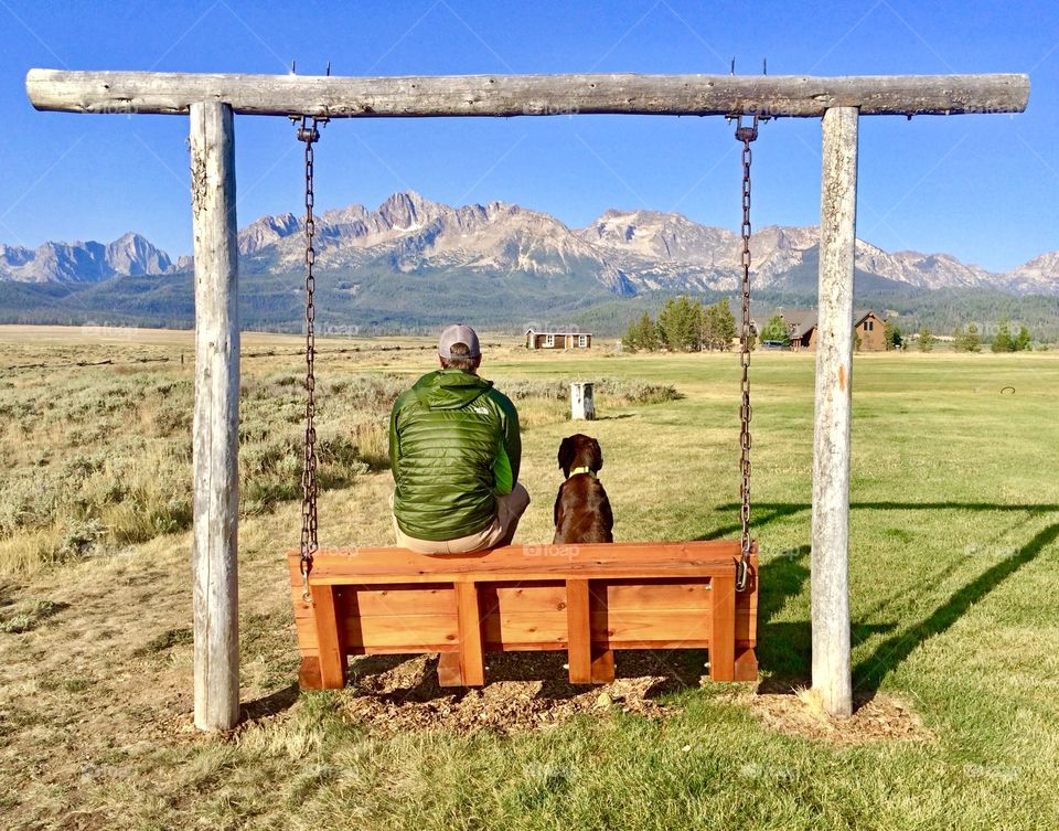 A man and his dog enjoying a relaxing spring day on a swing looking out on the Idaho Sawtooth mountain range.