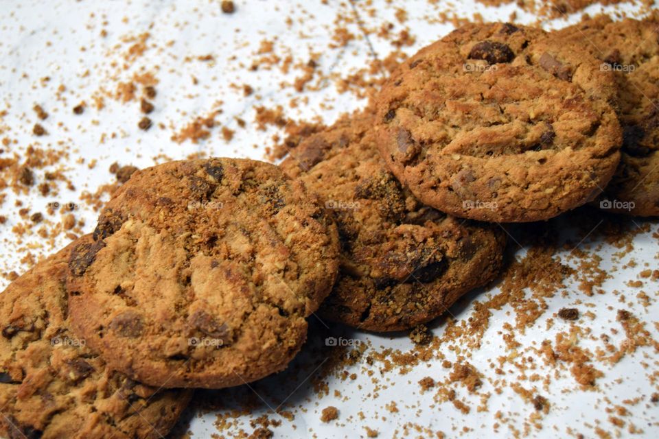 A diagonal row of delicious golden brown chocolate chip cookies laid across a white marbled background surrounded by chocolate chip cookie crumbs.