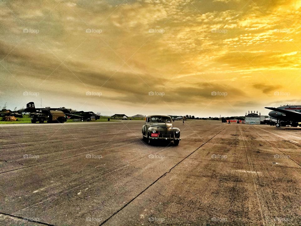 WWII Reenactment on airport runway, vintage car at sunset with war planes in back