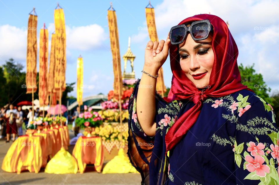 Portrait of a beautiful mother wearing a red hijab, wearing a floral patterned robe and wearing black glasses, taking a photo with a happy expression.