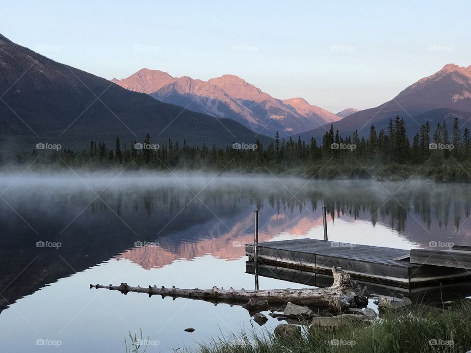Early sunrise over looking the lake with the Canadian Rockies in the background.  There is a log along the water’s edge next to a dock.  Steam is rising from the lake.
