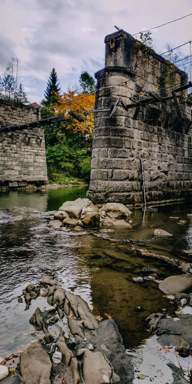 Old brick stone column of the bridge in the water. Transparent water in the mountain river, yellow green leaved trees. Stones in the water, beautiful nature location vertical view. Waterscape in Carpathian mountains. Brocken bridge, leaves in the wat