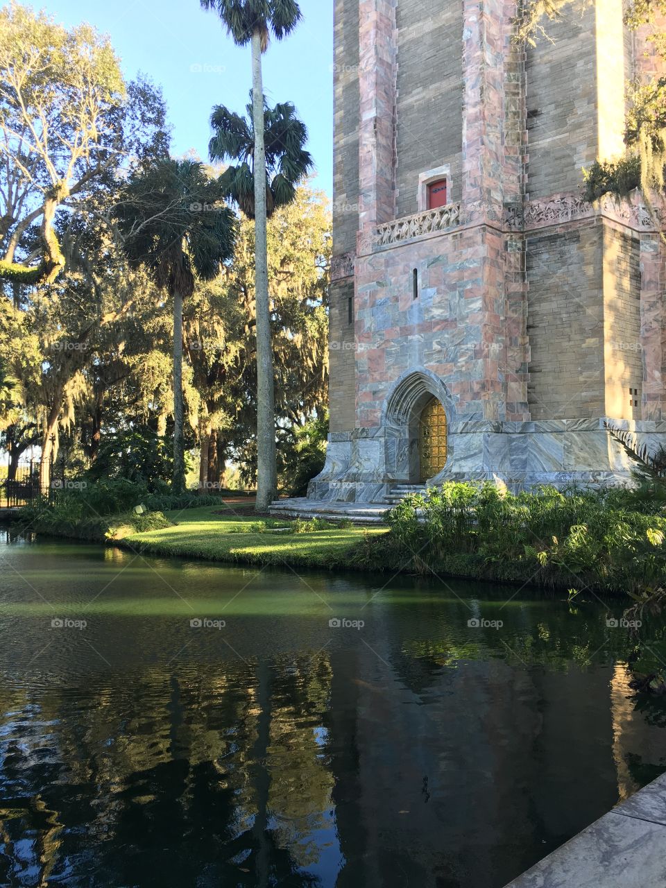 Tranquil scene of a pond lined with tropical trees and mossy live oaks near a tower with a golden door