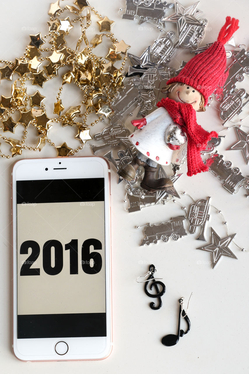 Overhead shot of a new year based scene. Iphone screen showing 2016 numbers, golden and silver coloured christmas ornamentsi a toy wearing a red hat and a red scarf. Treble clef shaped earring, musical note shaped black earring.