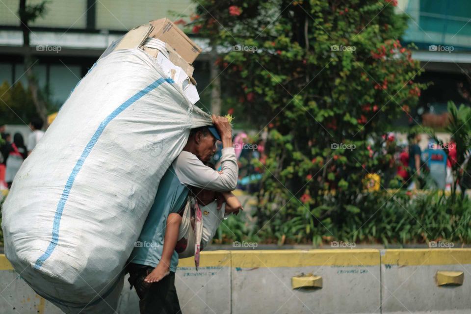 Old beggar carrying a huge sack on his back & a child on his front body. This picture taken on Jendral Sudirman Street during car free day; date: October 27, 2019.