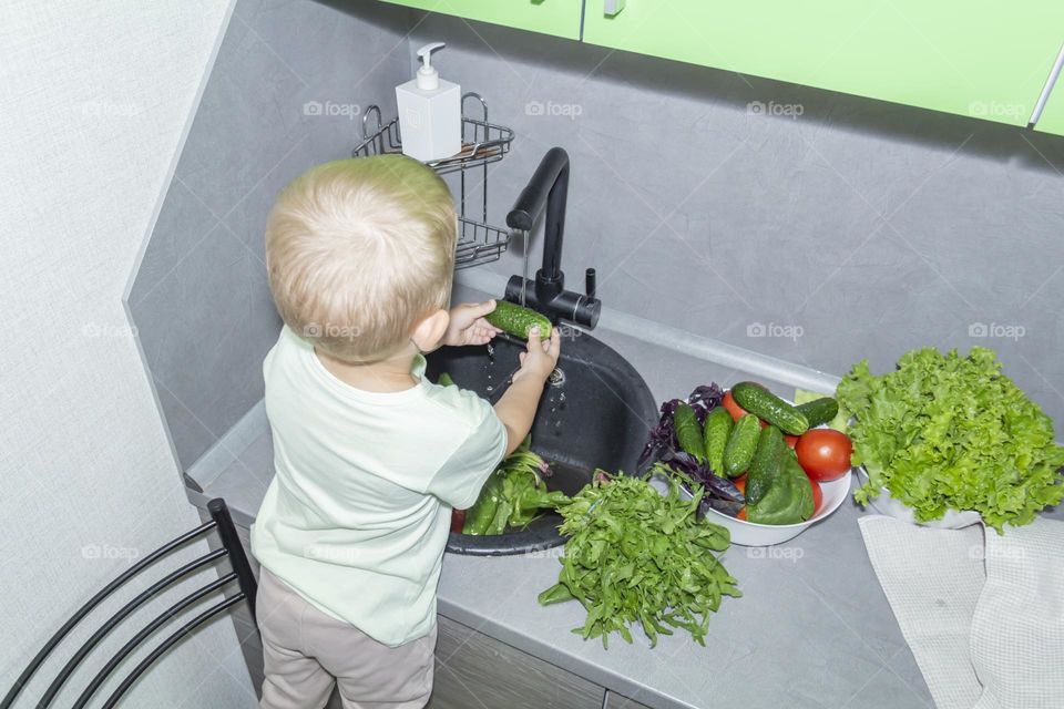 A small child stands on a chair near the sink and washes fresh vegetables under running tap water.