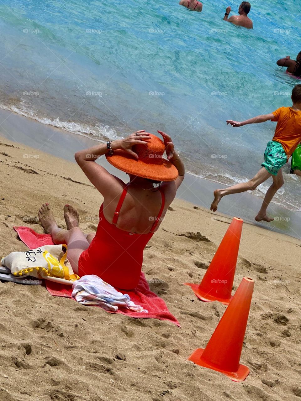 A woman in an orange suit with an orange hat on an orange towel prepares to enjoy the beach as a little kid runs by and people enjoy themselves in the water