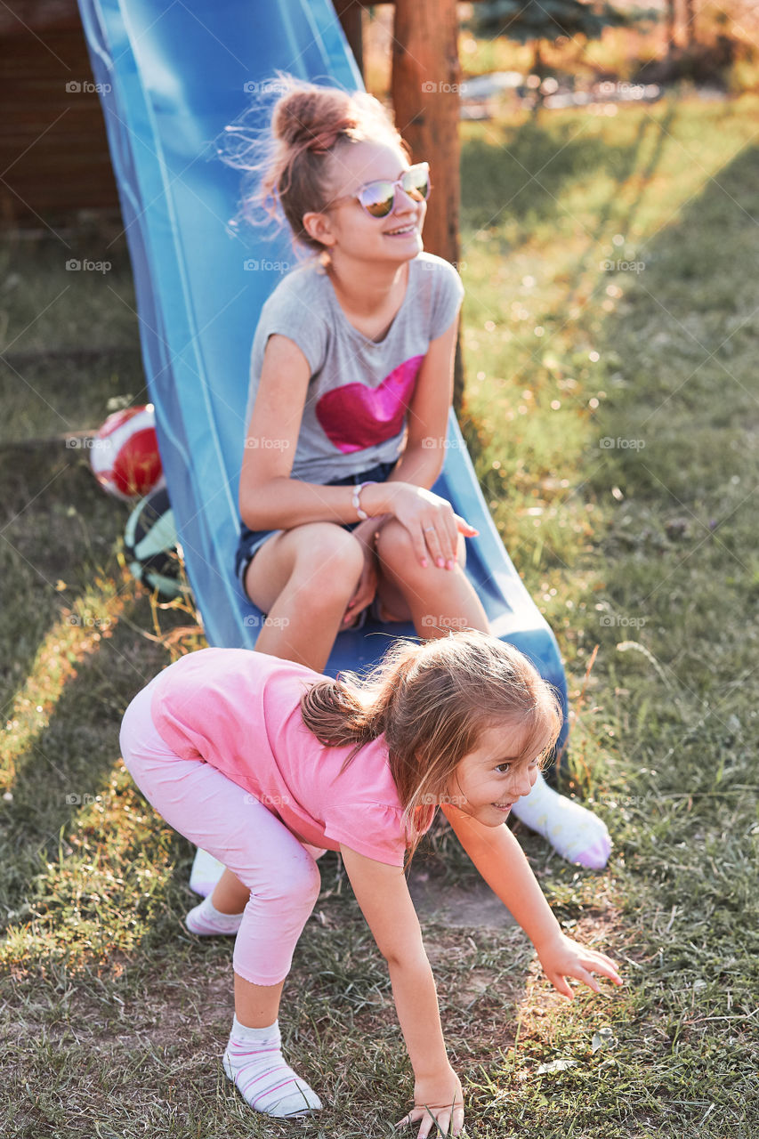 Teenage girl playing with her younger sister in a home playground in a backyard. Happy smiling sisters having fun on a slide together on summer day. Real people, authentic situations