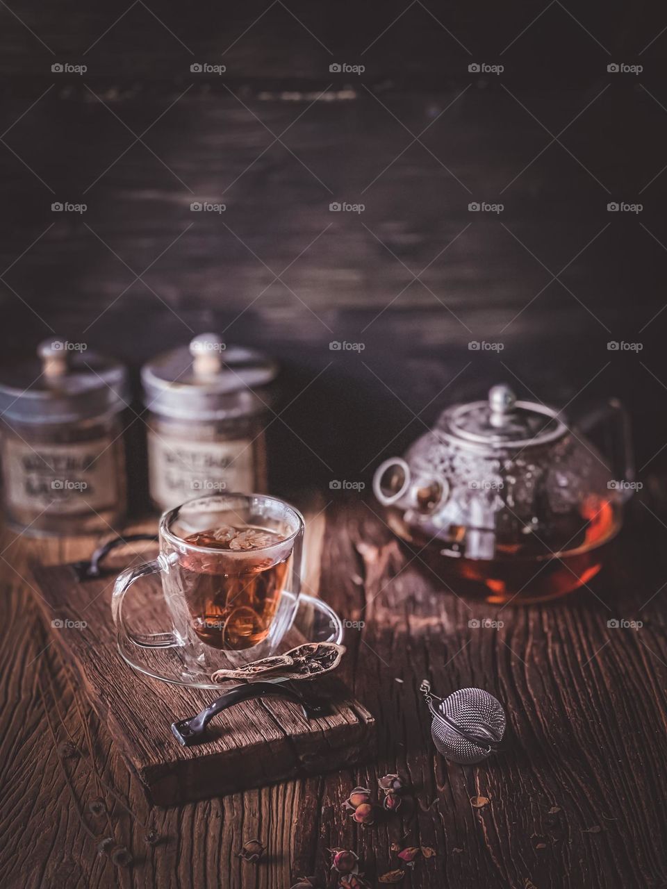 A cup of hot tea in a clear cup and saucer and a teapot on the side with dim lights and blurry background 