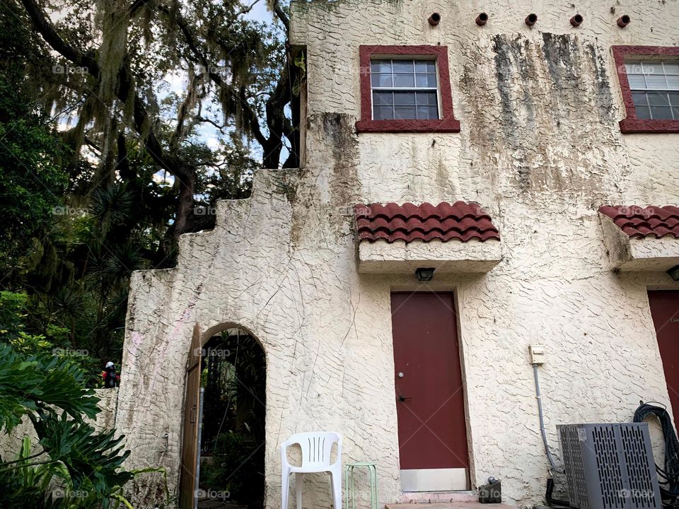 White and red spanish old style architecture residential large house built in the early 1900s. Doorway with antique style wood door in a stairway wall with white chair by a red door and window seen from tropical plants, live oak trees with child walk