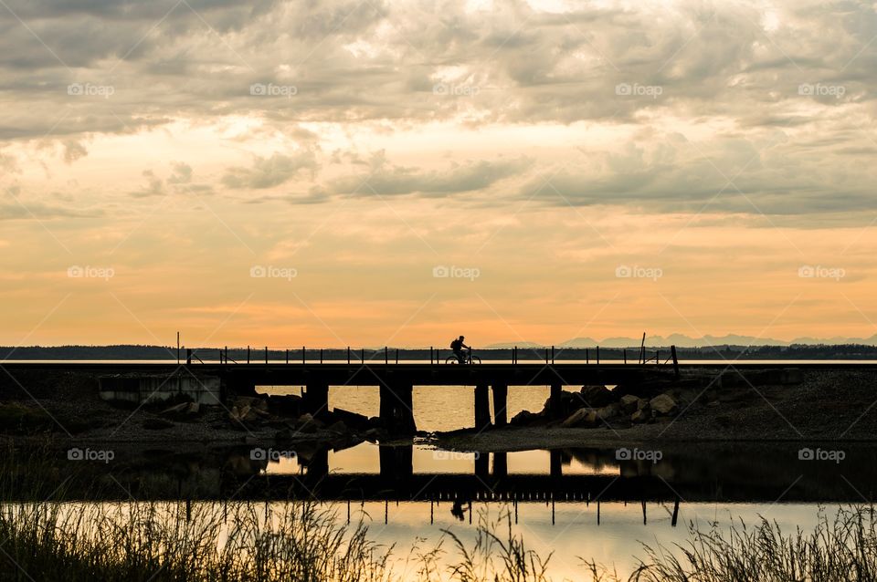 Biking on a trestle at sunset. Man bicycling on a railroad trestle over a pond on a cloudy sunset evening.