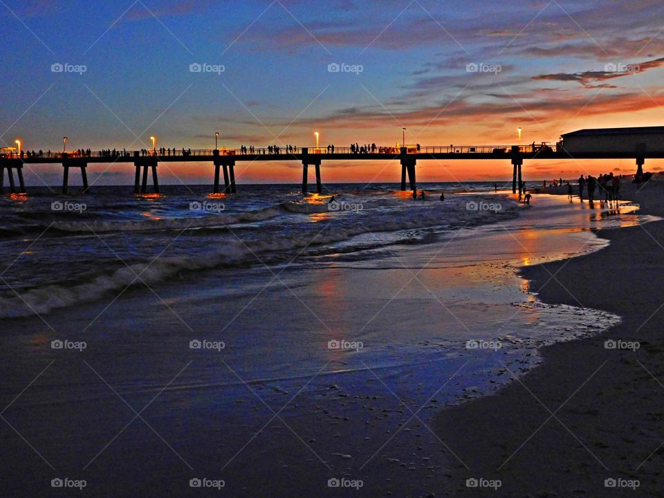 Shimmering sunset over the Gulf of Mexico as people walk along the sandy beach, swim and fish from the pier