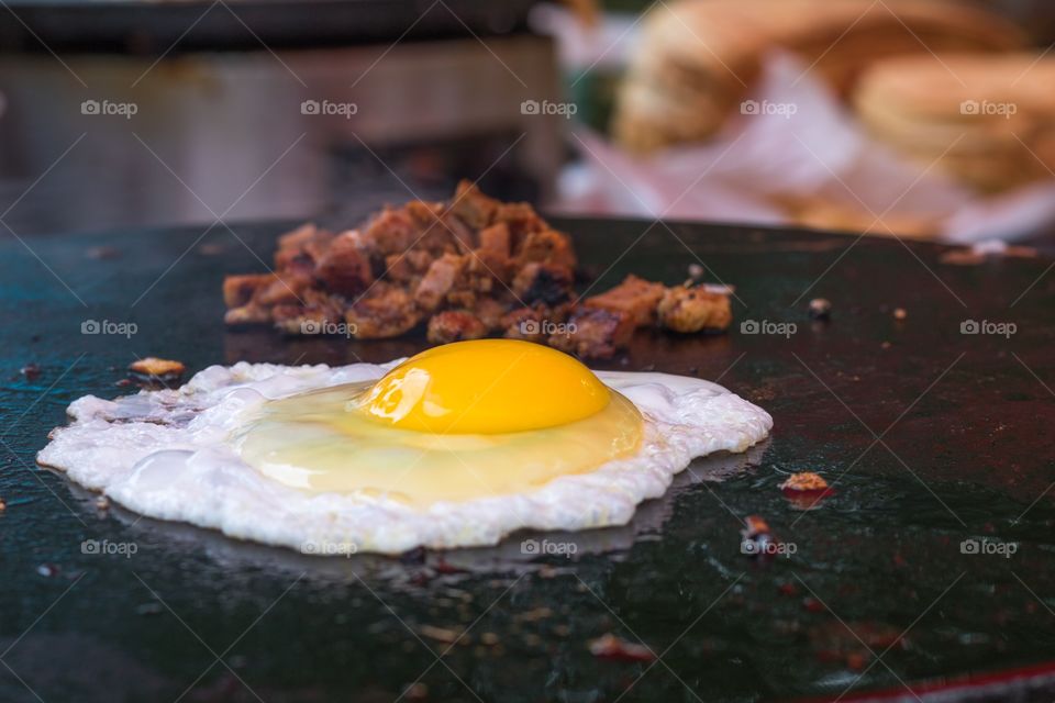 Closeup horizontal photo of an egg frying sunny side up on a round metal grill with a small mound of chopped bacon in soft focus behind it