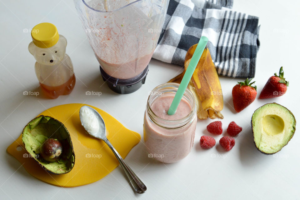 Fruit smoothie in a glass and ingredients consisting of a banana, raspberries, strawberries, avocado and honey next to a blender