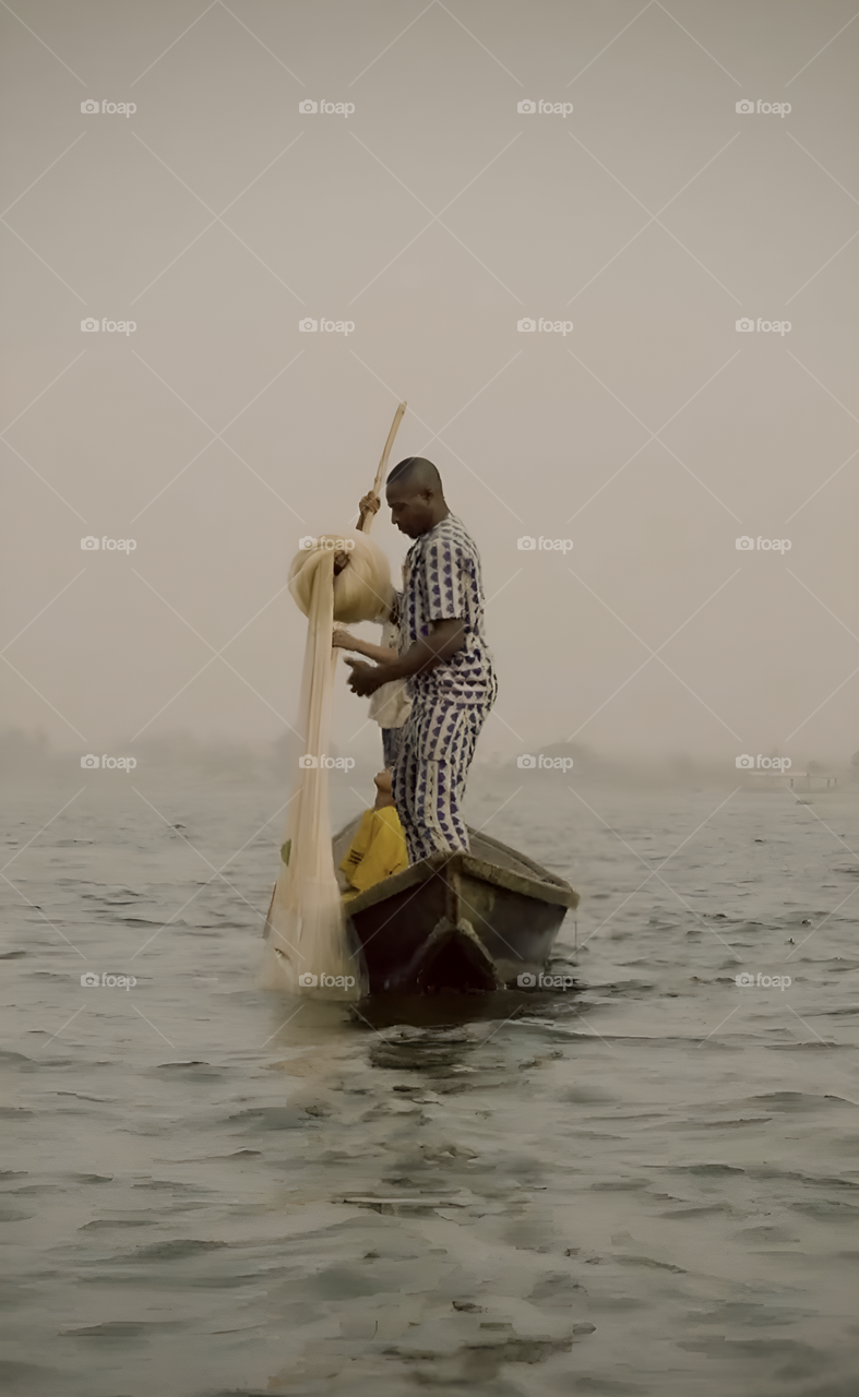 A fisherman stands on a wooden canoe, poised to cast his net into the shimmering water. His muscles tense with effort, the net spreads wide in mid-air. The tranquil backdrop of trees and sky adds serenity.