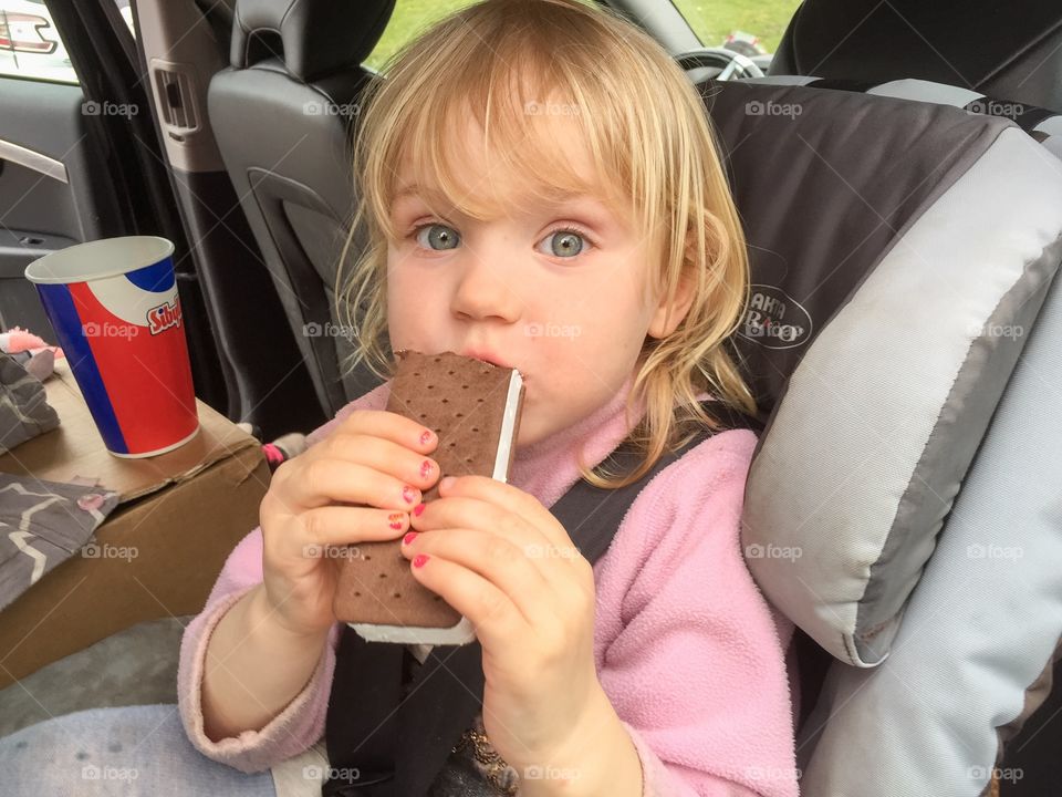 Three years old girl eating a sandwich ice cream in the car.