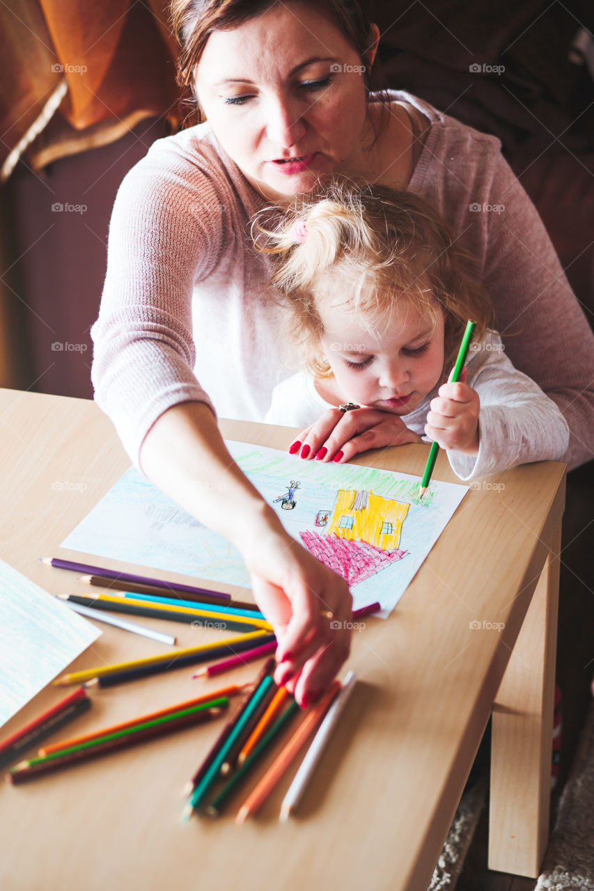 Mom with little daughter drawing a colorful pictures of house and playing children using pencil crayons sitting at table indoors