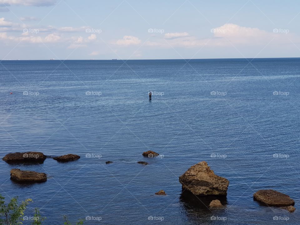 Lonesome person (a fisherman) standing in the sea; rocks on the foreground; blue water till the horizon.