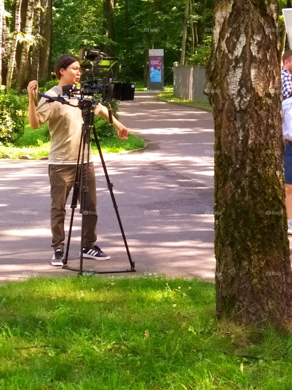 Summer day.  Filming a movie.  In the park, among the green trees, there is a man with a professional movie camera.  A man explains something and gesticulates