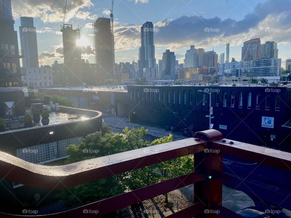 Over the railing of the outpost on top of the “Pulaski Bridge” at “Newtown Creek” we see the “BOXHOUSE Hotel” and beyond it the combined skylines of “Long Island City”, Queens and of “Manhattan”. 2023. Hypnotic Productions